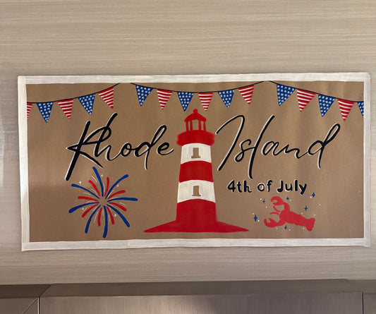 Decorative sign with a lighthouse, fireworks, and 'Rhode Island 4th of July' text on a brown background.
